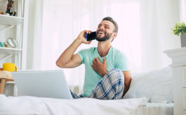 Gündelik giysiler içindeki yakışıklı genç adam yatakta otururken evde çalışıyor. Dizüstü bilgisayarı ve akıllı telefonu olan kendine güvenen bir adam yatak odasında kahve içer..