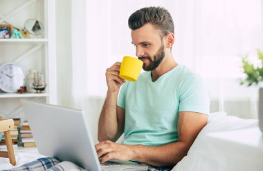 Gündelik giysiler içindeki yakışıklı genç adam yatakta otururken evde çalışıyor. Dizüstü bilgisayarı ve akıllı telefonu olan kendine güvenen bir adam yatak odasında kahve içer..