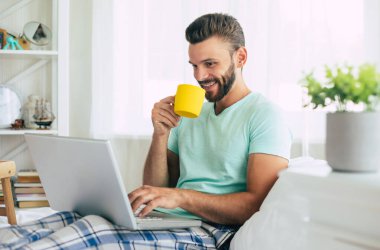 Gündelik giysiler içindeki yakışıklı genç adam yatakta otururken evde çalışıyor. Dizüstü bilgisayarı ve akıllı telefonu olan kendine güvenen bir adam yatak odasında kahve içer..