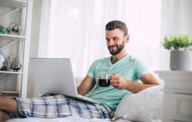 Gündelik giysiler içindeki yakışıklı genç adam yatakta otururken evde çalışıyor. Dizüstü bilgisayarı ve akıllı telefonu olan kendine güvenen bir adam yatak odasında kahve içer..