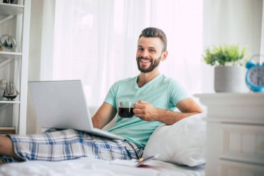 Gündelik giysiler içindeki yakışıklı genç adam yatakta otururken evde çalışıyor. Dizüstü bilgisayarı ve akıllı telefonu olan kendine güvenen bir adam yatak odasında kahve içer..