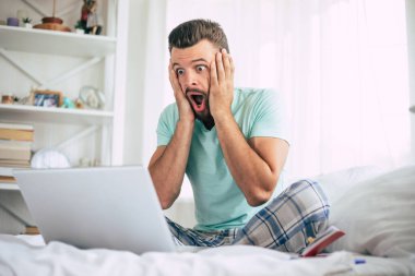 Heyecanlı, mutlu ve sakallı hayran evinde büyük beyaz bir yatakta dizüstü bilgisayarla kutlama yaparken bağırıyor.