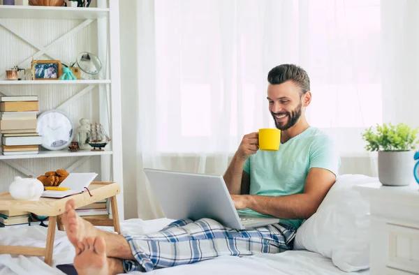 Gündelik giysiler içindeki yakışıklı genç adam yatakta otururken evde çalışıyor. Dizüstü bilgisayarı ve akıllı telefonu olan kendine güvenen bir adam yatak odasında kahve içer..