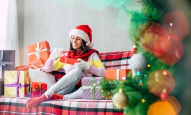 Kış tatilinin kutlaması. Noel Baba şapkalı ve kazaklı güzel, mutlu esmer kadın evdeki kanepede bir sürü hediye kutusuyla eğleniyor. Mutlu Noeller ve mutlu yıllar.