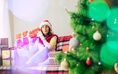 Kış tatilinin kutlaması. Noel Baba şapkalı ve kazaklı güzel, mutlu esmer kadın evdeki kanepede bir sürü hediye kutusuyla eğleniyor. Mutlu Noeller ve mutlu yıllar.