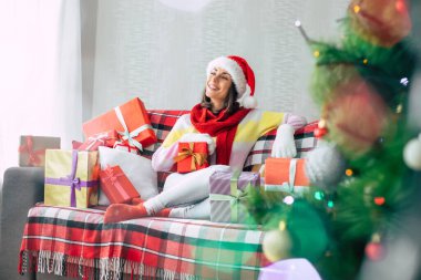 Kış tatilinin kutlaması. Noel Baba şapkalı ve kazaklı güzel, mutlu esmer kadın evdeki kanepede bir sürü hediye kutusuyla eğleniyor. Mutlu Noeller ve mutlu yıllar.