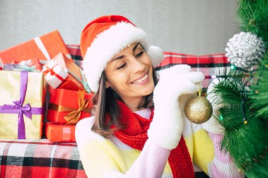 Süveter ve Noel Baba şapkalı güzel mutlu genç bir kadının yakın plan fotoğrafı hediye kutularının arkasında bir Noel ağacını süslüyor. Kış tatili