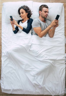 Güzel bir kadın ve yakışıklı genç adamın büyük beyaz bir yatakta yatarken akıllı telefon kullandıkları en iyi manzara fotoğrafı. İnternette sohbet ediyoruz. Çiftin üstündeki fotoğraf..