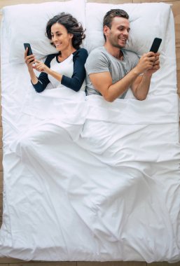 Güzel bir kadın ve yakışıklı genç adamın büyük beyaz bir yatakta yatarken akıllı telefon kullandıkları en iyi manzara fotoğrafı. İnternette sohbet ediyoruz. Çiftin üstündeki fotoğraf..