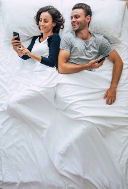 Güzel bir kadın ve yakışıklı genç adamın büyük beyaz bir yatakta yatarken akıllı telefon kullandıkları en iyi manzara fotoğrafı. İnternette sohbet ediyoruz. Çiftin üstündeki fotoğraf..