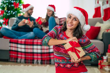Mutlu Noeller ve mutlu yıllar. Noel Baba şapkalı mutlu gülümseyen güzel bir kadının fotoğrafı bir grup arkadaş ve Noel ağacı arkaplanındaki hediye kutusunu gösteriyor..