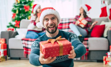 Mutlu Noeller ve mutlu yıllar. Noel Baba şapkalı mutlu sakallı genç adam elinde bir hediye kutusu, bir grup arkadaş ve Noel ağacı arkaplanı ile birlikte..