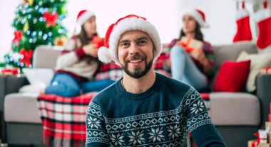 Mutlu Noeller ve mutlu yıllar. Noel Baba şapkalı mutlu sakallı genç adam elinde bir hediye kutusu, bir grup arkadaş ve Noel ağacı arkaplanı ile birlikte..