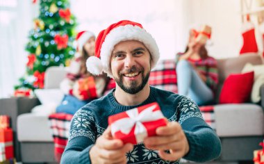 Mutlu Noeller ve mutlu yıllar. Noel Baba şapkalı mutlu sakallı genç adam elinde bir hediye kutusu, bir grup arkadaş ve Noel ağacı arkaplanı ile birlikte..
