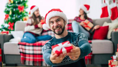 Mutlu Noeller ve mutlu yıllar. Noel Baba şapkalı mutlu sakallı genç adam elinde bir hediye kutusu, bir grup arkadaş ve Noel ağacı arkaplanı ile birlikte..