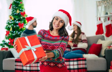 Mutlu Noeller ve mutlu yıllar. Noel Baba şapkalı mutlu gülümseyen güzel bir kadının fotoğrafı bir grup arkadaş ve Noel ağacı arkaplanındaki hediye kutusunu gösteriyor..
