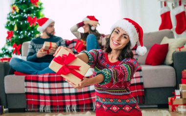Mutlu Noeller ve mutlu yıllar. Noel Baba şapkalı mutlu gülümseyen güzel bir kadının fotoğrafı bir grup arkadaş ve Noel ağacı arkaplanındaki hediye kutusunu gösteriyor..