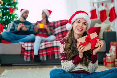 Noel süveteri ve Noel Baba şapkasıyla gülümseyen güzel genç bir kadın arkadaşlarının ve Noel ağacının arkasında elinde renkli bir hediye kutusu tutuyor..