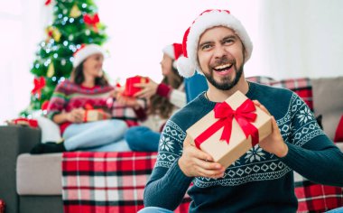 Noel Baba şapkası ve kışlık süveter giyen genç ve mutlu sakallı adam elinde hediye kutusuyla poz veriyor ve arka planda gülümseyen arkadaşlarıyla kamera önünde poz veriyor..