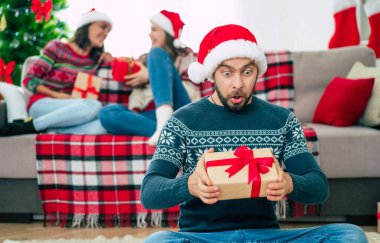 Noel Baba şapkası ve kışlık süveter giyen genç ve mutlu sakallı adam elinde hediye kutusuyla poz veriyor ve arka planda gülümseyen arkadaşlarıyla kamera önünde poz veriyor..