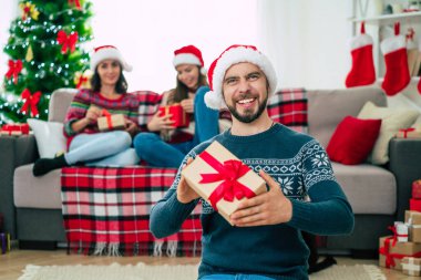Noel Baba şapkası ve kışlık süveter giyen genç ve mutlu sakallı adam elinde hediye kutusuyla poz veriyor ve arka planda gülümseyen arkadaşlarıyla kamera önünde poz veriyor..