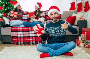 Noel Baba şapkası ve kışlık süveter giyen genç ve mutlu sakallı adam elinde hediye kutusuyla poz veriyor ve arka planda gülümseyen arkadaşlarıyla kamera önünde poz veriyor..