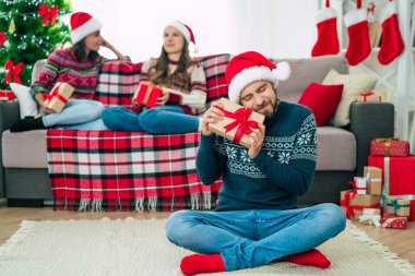 Noel Baba şapkası ve kışlık süveter giyen genç ve mutlu sakallı adam elinde hediye kutusuyla poz veriyor ve arka planda gülümseyen arkadaşlarıyla kamera önünde poz veriyor..