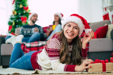 Noel süveteri ve Noel Baba şapkasıyla gülümseyen güzel genç bir kadın arkadaşlarının ve Noel ağacının arkasında elinde renkli bir hediye kutusu tutuyor..