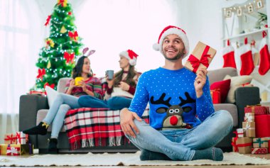 Noel hediyesi olan Noel Baba şapkalı yakışıklı genç adam poz veriyor ve evdeki mutlu arkadaşlarının arka planında kameraya bakıyor. 