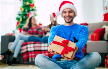 Noel hediyesi olan Noel Baba şapkalı yakışıklı genç adam poz veriyor ve evdeki mutlu arkadaşlarının arka planında kameraya bakıyor. 