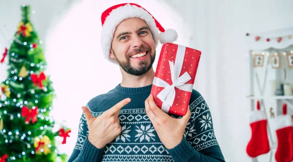 手の中にクリスマスの贈り物とサンタの帽子のハンサムな若いひげを生やした笑顔の男の肖像画を閉じてカメラでそれを示しています — ストック写真