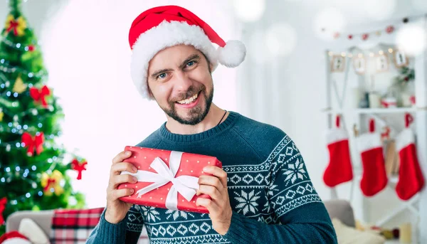 手の中にクリスマスの贈り物とサンタの帽子のハンサムな若いひげを生やした笑顔の男の肖像画を閉じてカメラでそれを示しています — ストック写真