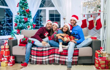 Noel Baba şapkalı ve elinde hediyeler olan bir grup güzel arkadaş Noel ağacı arka planında poz veriyor ve eğleniyorlar.