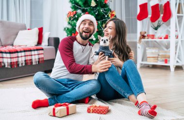 Noel romantik hediyesi. Mutlu, birbirine aşık genç bir çift Noel ağacı arka planında yerde otururken birbirlerine sürpriz yapıp hediyeler veriyorlar..