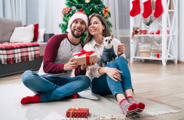 Noel romantik hediyesi. Mutlu, birbirine aşık genç bir çift Noel ağacı arka planında yerde otururken birbirlerine sürpriz yapıp hediyeler veriyorlar..