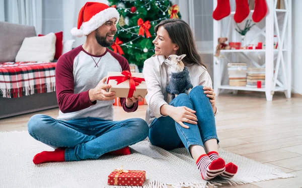 Noel romantik hediyesi. Mutlu, birbirine aşık genç bir çift Noel ağacı arka planında yerde otururken birbirlerine sürpriz yapıp hediyeler veriyorlar..