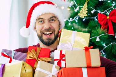 Noel Baba şapkalı yakışıklı, sakallı ve elinde hediyeler olan genç adam Noel ağacı arka planında poz veriyor.