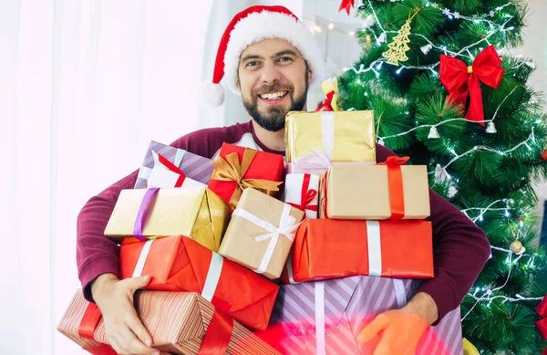 ハンサム励起ひげを生やした若いです男でサンタ帽子で多くのプレゼントで手は上のクリスマスツリーの背景 — ストック写真