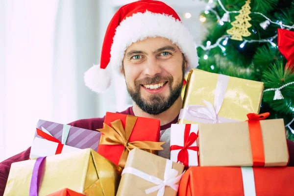 ハンサム励起ひげを生やした若いです男でサンタ帽子で多くのプレゼントで手は上のクリスマスツリーの背景 — ストック写真