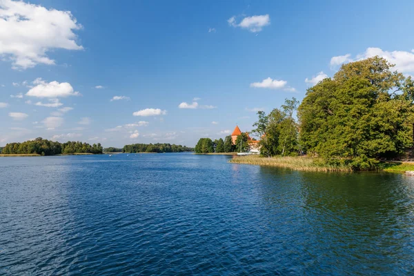 Scenic lake view with clear blue water, green trees, and medieval castle tower in the distance under blue sky with clouds