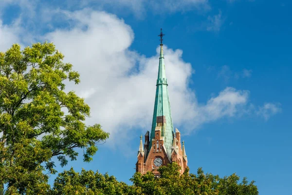 Historic church tower with copper spire and clock, rising above green trees under blue sky with clouds, perfect for architecture, travel, religion and cultural heritage projects