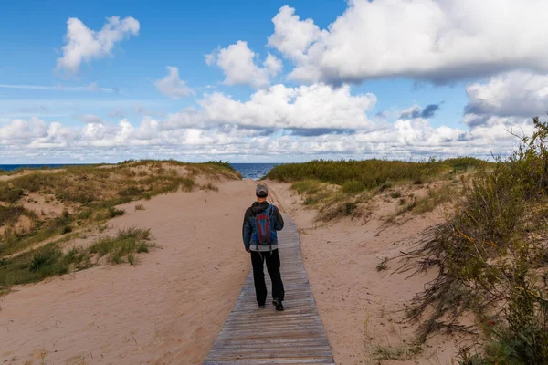 Erkek yürüyüşçü, kum tepelerinden kumsala doğru yürüyor kümülüs bulutlarıyla mavi gökyüzünün altında, huzurlu kıyı manzarası, seyahat, doğa ve açık hava macera projeleri için mükemmel.