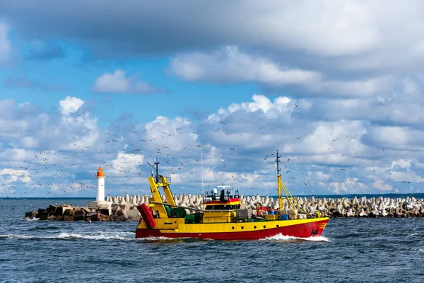 Fishing boat returns home from sea to harbor surrounded by seagulls, with lighthouse in background under dramatic sky, perfect for travel, maritime, fishing industry and coastal projects