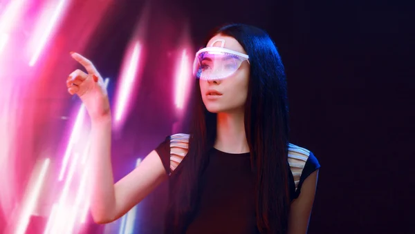 Beautiful woman in futuristic dress over dark background. Gamer girl in glasses of virtual reality with controller in hands. Augmented reality, game, future technology, hobby concept. VR.