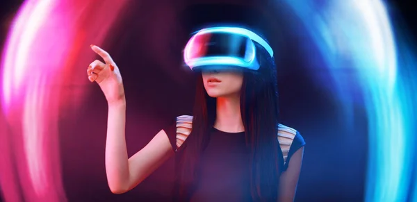 Beautiful woman in futuristic dress over dark background. Gamer girl in glasses of virtual reality with controller in hands. Augmented reality, game, future technology, hobby concept. VR.