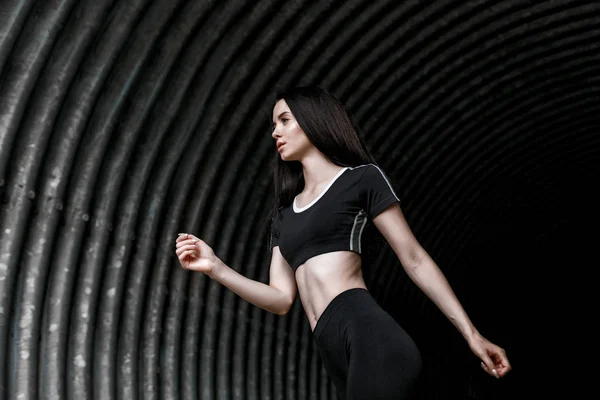 Sports woman running in fashion sportswear on tunnel urban gray background. Fitness model working out outdoor. Young beautiful slim brunette girl in trendy black leggings and top.