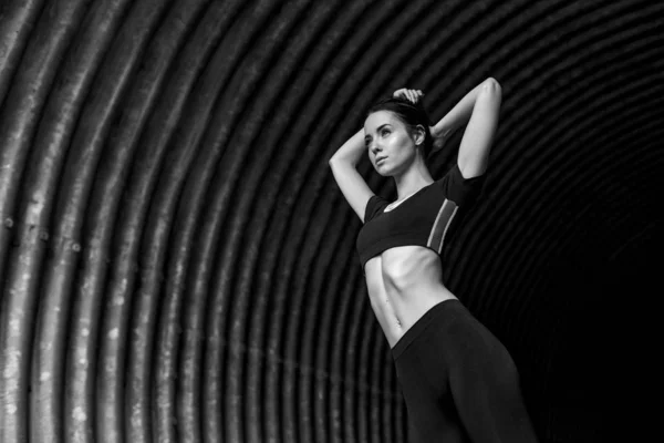 Sports woman posing in fashion sportswear on tunnel urban background. Fitness model working out outdoor. Young beautiful slim brunette girl in trendy leggings and top. Black and white.