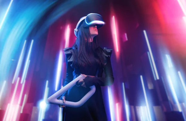 Beautiful woman in futuristic dress over dark background. Gamer girl in glasses of virtual reality with controller in hands. Augmented reality, game, future technology, hobby concept. VR.