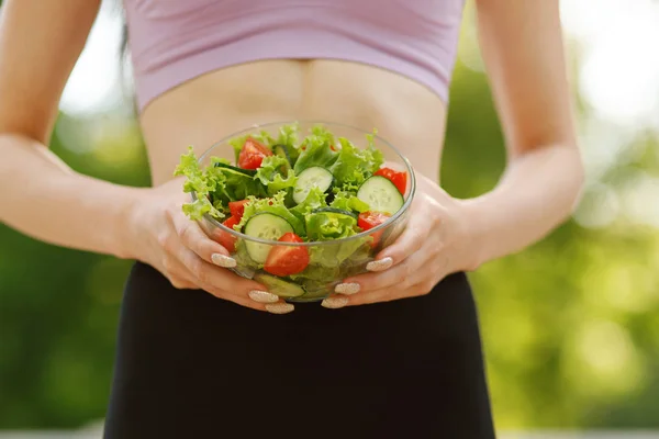 Sports woman holding a bowl with fresh salad at the park. Young beautiful slim brunette girl outdoor