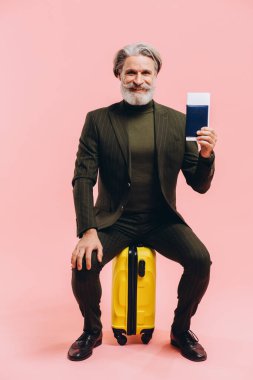 Takım elbiseli şık orta yaşlı bir adam sarı bir bavula oturur ve bir pasaport tutar..